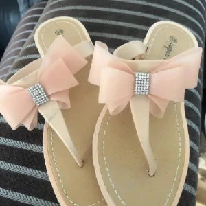 Beiga/rosa sandaler med rosett och strass - Snygga beiga sandaler med stor ljusrosa rosett och glittrande strassdetalj framtill. Perfekta för sommaren med öppen tå och platt sula. Mjukt material och stilren design som sticker ut.