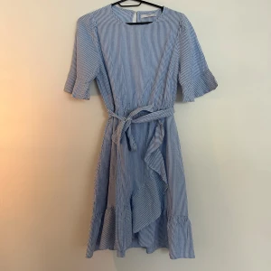 Blåvit randig omlottklänning med volang - Superfin ljusblå och vit randig klänning med omlottkänsla, volangdetaljer och knytband i midjan. Jätte fin. Aldrig använd 