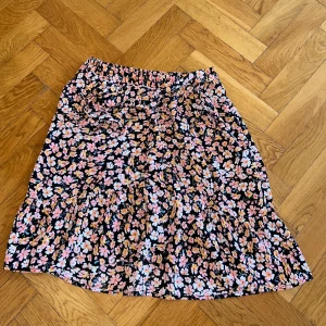 Blommig kortkjol med volang - Supersöt kortkjol med blommigt mönster i rosa, vitt och orange på svart botten. Kjolen har volang nertill och resår i midjan för skön passform även knyte då man själv kan välja passform.  