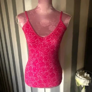 Rosa blommig linnetopp med smala band - Snygg rosa linnetopp med blommigt mönster och smala axelband. Toppen har en v-ringad hals och sitter tight mot kroppen. Perfekt för dig som gillar färg och vill sticka ut lite extra i sommarvärmen. Med inbyggd bh. Storlek M på lappen men lite mindre 