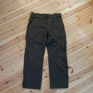 Mörkgröna cargopants med fickor - Säljer ett par mörkgröna cargopants med klassiska benfickor och avslappnad passform. Byxorna har knapp och dragkedja framtill samt bälteshällor. Perfekta för en chill och funktionell stil.