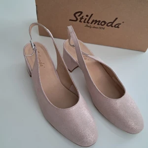 Glittriga ljusrosa pumps från Stilmoda - Snygga ljusrosa pumps från Stilmoda med glittrig yta och öppen häl. Skorna har rund tå, låg klack och med spänne vid ankeln. Perfekta för dig som vill ha en chic och trendig look med italiensk känsla.