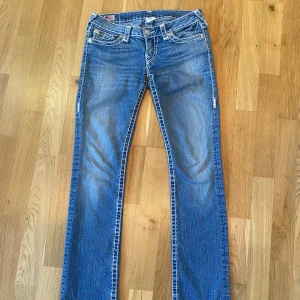 True Religion blå bootcut jeans - Säljer ett par blå jeans från True Religion med tydliga vita kontrastsömmar och ikoniska bakfickor. Modellen har bootcut-ben och låg midja. Klassisk femficksdesign och snygg tvättad denim. Perfekta för dig som gillar statement-jeans med coola detaljer.