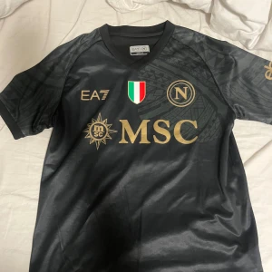 Ea7 armani Napoli fotbollströja - Snygg svart Napoli fotbollströja från EA7 med guldiga detaljer och loggor. Tröjan är oanvänd