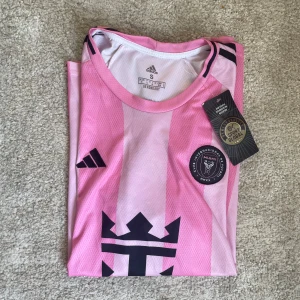 Inter Miami Messi 10 Adidas fotbollströja - Säljer en officiell Inter Miami fotbollströja från Adidas i rosa med svarta detaljer. Tröjan har Messi 10-tryck på ryggen, klubbmärke på bröstet och klassiska Adidas-ränder på axlarna. Tillverkad i lätt och ventilerande AEROREADY-material, perfekt för match eller träning.Hör av dig vid frågor, pris kan diskuteras ❤️