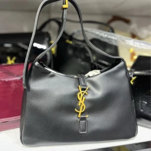 Svart YSL axelväska i skinn - Snygg svart axelväska från YSL i slätt skinn med guldigt YSL-logo framtill. Väskan har en enkel, stilren design med justerbart axelband och dragkedja upptill. Perfekt storlek för det viktigaste och insida med fack för småsaker.