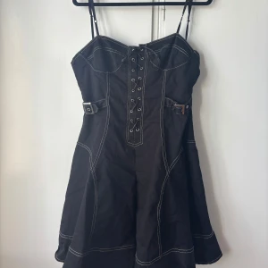 Svart kort klänning med snörning - Svart kort klänning med smala axelband, markerade sömmar och snörning framtill. Klänningen har coola bältesdetaljer i midjan och en lätt utsvängd kjol. Perfekt för dig som gillar edgy och unik stil. Hör av dig om du vill ha fler bilder eller har några frågor! 