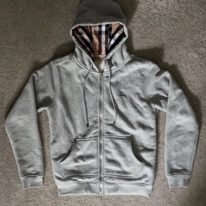 Grå Burberry Zip - Säljer en riktigt fet grå zip från Burberry med klassiskt rutigt foder i huvan och längs insidan. Tröjan har dragkedja, snörning i huvan, broderad logga på bröstet och två stora fickor framtill. Skick 10/10. Hör gärna av er vid funderingar, Mvh