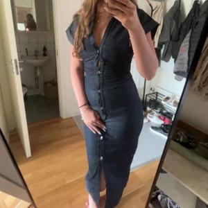 Mörkblå jeansklänning med knappar - Säljer en mörkblå långklänning i jeansmaterial med korta ärmar och snygg v-ringning. Klänningen har knappar hela vägen framtill och en liten slits nedtill. Sitter figurnära och har en modern, stilren look. Lappen kvar 💕inga defekter 💕 säljer endast då den var lite liten på mig