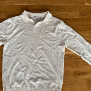 Vit stickad pikétröja från Sandö - Klassisk vit pikétröja från Sandö i stickad bomull. Tröjan har krage och v-ringning, samt långa ärmar och ribbade muddar. Perfekt för en clean och stilren look, funkar lika bra till jeans som till chinos.