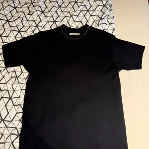 Svart t-shirt från Acne Studios - Svart t-shirt från Acne Studios med diskret logga broderad runt halsringningen. Klassisk passform och korta ärmar, perfekt för en clean och stilren look. Tillverkad i mjuk bomull som känns skön mot huden. Aldrig använt!