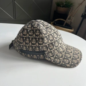 Salvatore Ferragamo mönstrad keps - Snygg keps från Salvatore Ferragamo med beige och svart mönster över hela kepsen. Klassisk böjd skärm och justerbar rem bak. Tillverkad i Italien och har exklusiv känsla med tydlig logga i mönstret. Perfekt accessoar för att lyfta din stil.