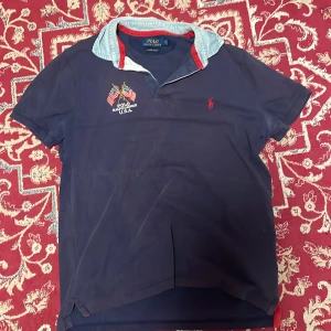 Mörkblå pikétröja Polo Ralph Lauren - Snygg mörkblå pikétröja från Polo Ralph Lauren med ljusblå krage och röda detaljer. Broderade amerikanska flaggor och logga på bröstet. Klassisk passform och korta ärmar, perfekt för en avslappnad men stilren look.