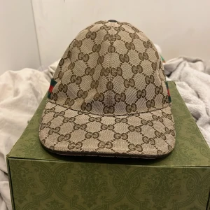 Gucci keps med GG-mönster och rödgrön detalj - Snygg keps från Gucci i beige och brunt med klassiskt GG-monogram över hela kepsen. Kepsen har en böjd skärm och justerbar rem baktill i mörkbrunt skinn. Sidorna pryds av Guccis ikoniska rödgröna band. Perfekt för dig som vill sticka ut med lyxig streetstyle.