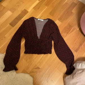 Blommig blus med v-ringning från NA-KD - Supersöt blommig blus från NA-KD i svart med små röda blommor. Djup v-ringning, långa puffiga ärmar och smockad midja som ger en croppad look. Perfekt för dig som gillar romantiska detaljer och vill sticka ut lite extra.