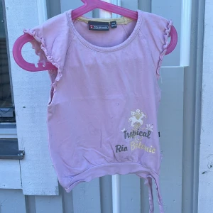 Ljusrosa topp med volang från Color Kids - Supersöt ljusrosa topp från Color Kids med volangdetaljer på ärmarna och tryck med texten 'Tropical Rio Botanic' i guld och silver. Toppen har knytband nertill och är ärmlös, perfekt för varma dagar.