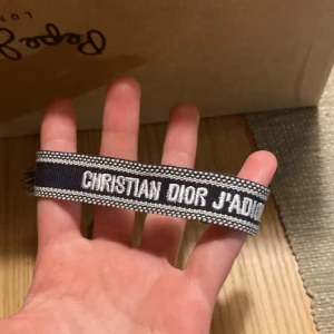 Christian Dior J'adior armband - Snyggt armband från Christian Dior med texten 'J'adior' broderad i vitt på mörkblå bakgrund. Armbandet har justerbar knytning med tofsar i ändarna och är gjort i ett mjukt textilmaterial. Perfekt accessoar för att lyfta din outfit.