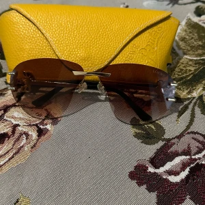 Loewe solglasögon med bruna glas - Snygga solglasögon från Loewe med bruna tonade glas och guldfärgade detaljer. Glasögonen har en modern aviatorform och tunna metallskalmar med Loewe-logga på sidorna. Kommer med ett gult fodral i läder-look. Perfekt accessoar för att lyfta din stil.