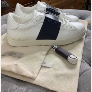 Valentino Open sneakers vit/svart - Snygga Valentino Open sneakers i vitt skinn med bred svart rem över mitten. Klassisk låg modell med rund tå och platt sula. Ikoniska nitar baktill på sulan ger extra edge. Perfekta för dig som gillar stilrena och exklusiva sneakers.