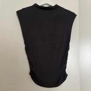 Svart ärmlös topp från Gina Tricot - Svart ärmlös topp från Gina Tricot med hög hals och snygga rynkade sidor. Toppens material är mjukt och stretchigt, perfekt för en trendig och clean look. Passformen är normal och modellen är enkel men ändå modern. Finns deo fläckar som går bort enkelt se bilderna. Säljer även den matchande kjolen.