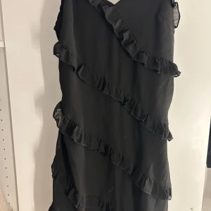 Svart volangklänning med smala axelband - Supersöt svart klänning med smala axelband och flera diagonala volangdetaljer över hela klänningen. Lätt och flowy känsla, perfekt för dig som gillar en enkel men ändå lekfull stil. V-ringad framtill och justerbara axelband för bästa passform. Aldrig använd 