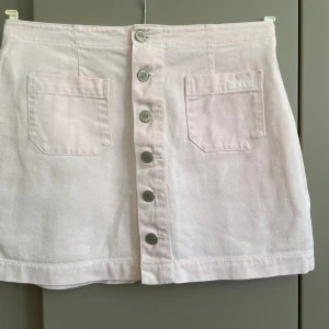 Ljusrosa jeanskjol från Bongo - Supersöt ljusrosa kort jeanskjol från Bongo med raka linjer, två stora fickor fram och snygga silverfärgade knappar hela vägen. Perfekt för dig som gillar en clean och enkel look med en touch av retro. Midjemått 78 cm