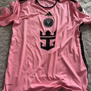 Inter Miami Messi 10 rosa matchtröja - Inter Miami CF fotbollströja i rosa med svarta detaljer, Messi 10 på ryggen och klubbmärke på bröstet. Tillverkad i lätt och ventilerande material med AEROREADY-teknologi. Klassisk Adidas-design med svarta ränder på axlarna och MLS-logga på ärmen.