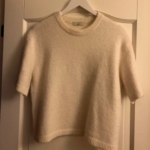 Krämvit stickad tröj - Superfin krämvit stickad tröja från Filippa K med korta ärmar och rund hals. Tröjan har en mjuk och lite boxig passform, perfekt att styla med jeans eller kjol. Ribbstickade detaljer vid ärmslut, hals och nederkant ger en clean look.