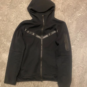 Nike Tech Fleece  - Säljer denna svarta Tech Fleece från Nike med hel dragkedja framtill och huva. Snygga svarta detaljer i form av ett V-format band över bröstet och en extra ficka med dragkedja på ärmen. Perfekt för dig som gillar sportig och clean stil. Köpt i Thailand, Storlek L motsvarar M i Europa.