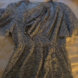 Blommig blå omlottklänning - Supersöt kort omlottklänning i blått med vitt blommönster. Klänningen har korta ärmar och knyt i midjan för en snygg passform. Perfekt för varma dagar och ger en fräsch sommarkänsla.