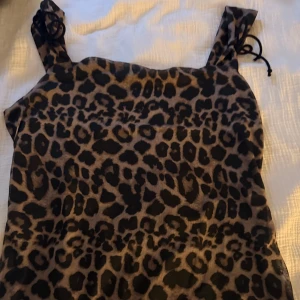 Leopardmönstrad kort klänning - Snygg kort klänning med leopardmönster och breda axelband med knyt. Klänningen har en figurnära passform och är perfekt för dig som vill sticka ut. Färgerna går i brunt och svart, vilket ger en cool och trendig look.