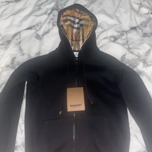 Svart Burberry hoodie med klassisk huva - Svart hoodie från Burberry i storlek M med dragkedja och huva fodrad i det ikoniska rutiga Burberry-mönstret. Tröjan har snörning i huvan och praktiska fickor framtill. Lite mindre i storlek, om du är mellan s/m passar den.