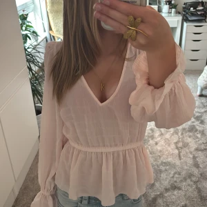 Ljusrosa blus med volang och v-ringning - Supersöt ljusrosa blus från Gina Tricot med v-ringning, långa puffiga ärmar och volang nedtill. Endast använd ett fåtal gånger.