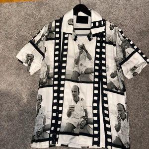 Svartvit skjorta från Limitato - Unik svartvit skjorta från Limitato med filmrulle-mönster och fotoprint av en man i retrostil. Skjortan har korta ärmar, klassisk krage och knäppning framtill. Nypris: 1899kr.    skick: 10/10. Strl M