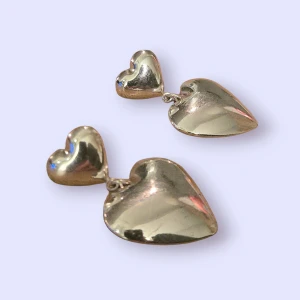 Silverfärgade hjärtformade örhängen - Säljer ett par snygga örhängen i silverfärg med dubbla hjärtan. De har en blank, spegelliknande yta och är riktigt statement med sin coola, hängande design. Perfekta för dig som vill sticka ut och addera lite extra kärlek till din stil.
