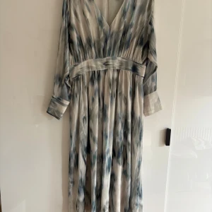 Långklänning från H&M med blå-grått mönster - Säljer en långklänning från H&M i storlek L. Klänningen har ett abstrakt blått-grått mönster, v-ringning och långa ärmar med breda manschetter. Materialet är mjukt och har ett fint fall. Använd en gång. 