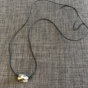 Halsband med färgglad pärla - Unikt halsband med svart läderrem och en stor, handgjord pärla i vitt med röda och blå detaljer. Pärlan har ett coolt, nästan bohemiskt mönster och ger halsbandet en personlig touch. Perfekt för dig som gillar att sticka ut med accessoarer. Hör av dig om du vill ha fler bilder eller har några funderingar! 