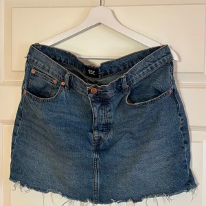 Blå jeanskjol från 157 Maddy, stl L - Snygg blå jeanskjol från 157 Maddy i storlek L. Klassisk femficksmodell med rå, fransig kant nertill för en cool vibe. Kjolen har normal passform och är perfekt till sneakers eller boots. Enkel att matcha med både t-shirt och hoodie.