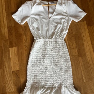 Vit klänning från Bubbleroom - Superfin vit klänning från Bubbleroom med v-ringning, korta ärmar och smockad midja och kjol. Klänningen har volangdetalj nertill och är tillverkad i ett lätt, glansigt material som känns lyxigt. Perfekt för sommarens alla tillfällen! Nypris 800kr. Säljs för 149kr.