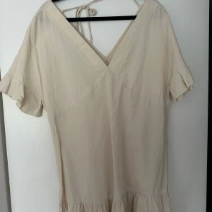 Beige kortklänning med volangärm - Supersöt beige kortklänning med v-ringning och knyt i nacken. Klänningen har volangdetaljer på ärmarna och nertill, samt korta ärmar. Perfekt för varma dagar och enkel att styla med sneakers eller sandaler.