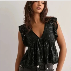 Svart glittrig volangtopp - Supersnygg svart blus med glittriga paljetter och volangdetaljer vid axlarna. V-ringning och luftig. Från Nelly, helt slutsåld. Använd en gång. 