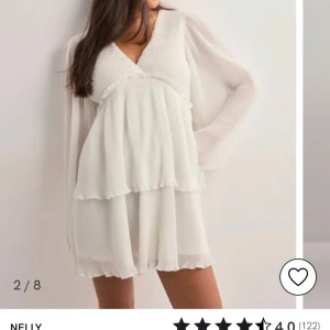 Vit omlottklänning från Nelly - Supersöt vit kortklänning från Nelly med v-ringning, omlottliv och långa, transparenta ärmar. Klänningen har volangdetaljer och ett luftigt, plisserat tyg som ger en romantisk vibe. Perfekt för sommarens alla tillfällen!