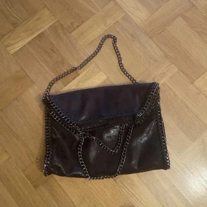 Stella McCartney liknande väska - Cool svart axelväska med glansig finish och kraftiga silverfärgade kedjor längs kanterna och som axelrem. Väskan har en envelope-formad front och är perfekt för dig som gillar en edgy stil. Materialet ser ut som syntet med skinnlook. LIKNANDE VÄSKA 