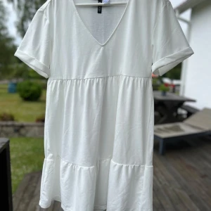 Vit kortklänning med v-ringning - Superfin vit kortklänning från Gina Tricot med v-ringad hals och korta ärmar. Klänningen har en avslappnad passform och volangdetaljer nertill, perfekt för en fräsch och enkel look. Tillverkad i mjuk bomull som känns skön mot huden.