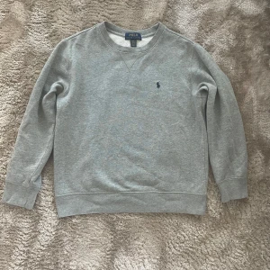 Grå sweatshirt från Polo Ralph Lauren - Klassisk grå sweatshirt från Polo Ralph Lauren med rund halsringning och broderad marinblå logga på bröstet. Tröjan har långa ärmar och ribbade muddar vid ärmslut och nederkant. I felfritt skick! 