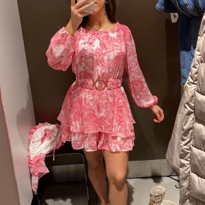 Rosa blommig jumpsuit med volang med tag kvar - Superfin oanvänd jumpsuit korall-rosa och vit med blommigt mönster, volangkjol och markerad midja med snyggt bälte. Långa ärmar och kan bäras offshoulder. Perfekt för sommarens alla tillfällen! Nypris 299, säljer för 180kr