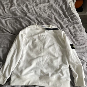 Vit sweatshirt från Stone Island - Säljer en clean vit sweatshirt från Stone Island med klassisk logga på ärmen. Tröjan har rund hals, ribbade muddar och är tillverkad i mjuk bomull. Perfekt för dig som gillar stilrena och exklusiva plagg med streetkänsla.