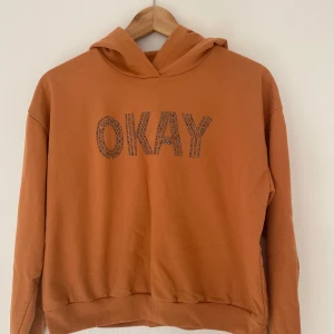 Orange hoodie från Lindex -  orange hoodie från Lindex med huva och texten 'OKAY' framtill i glittriga strassdetaljer. Mjuk insida och ribbade muddar. Perfekt för dig som gillar en chill och trendig stil.