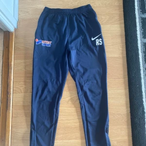 Nike Dri-Fit träningsbyxor marinblå L - Marinblå träningsbyxor från Nike med Dri-Fit-teknologi, resår i midjan och dragkedjor vid bensluten. Byxorna har tryck med Intersport-logga och initialerna RS på ena benet. Perfekta för fotboll eller annan träning.