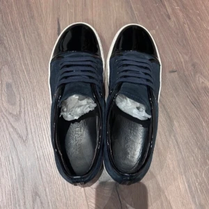 Svarta och blå sneakers från Lanvin - Snygga sneakers från Lanvin med en kombination av svart lack och blå mocka. Skorna har en vit sula och klassisk snörning. Perfekta för en stilren look. byten tillgänglig.